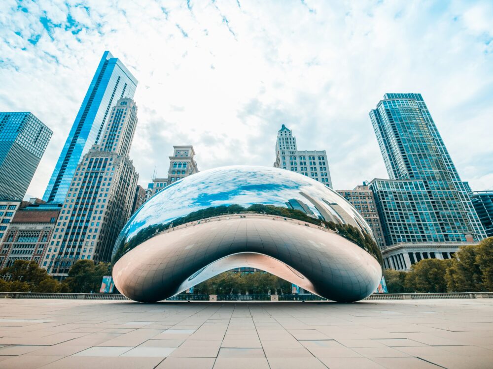 A large, reflective sculpture known as The Bean sits in an open plaza, surrounded by modern skyscrapers and trees under a partly cloudy sky in an urban cityscape.