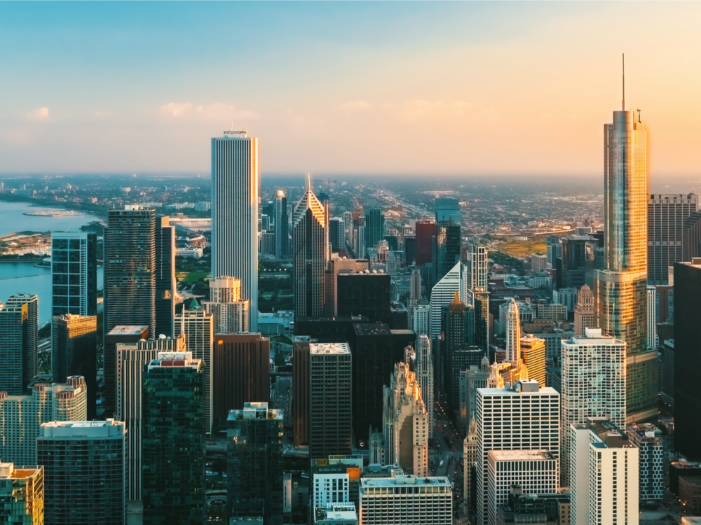 Aerial view of a city skyline with tall skyscrapers and buildings at sunset, a river on the left, and a clear sky above.