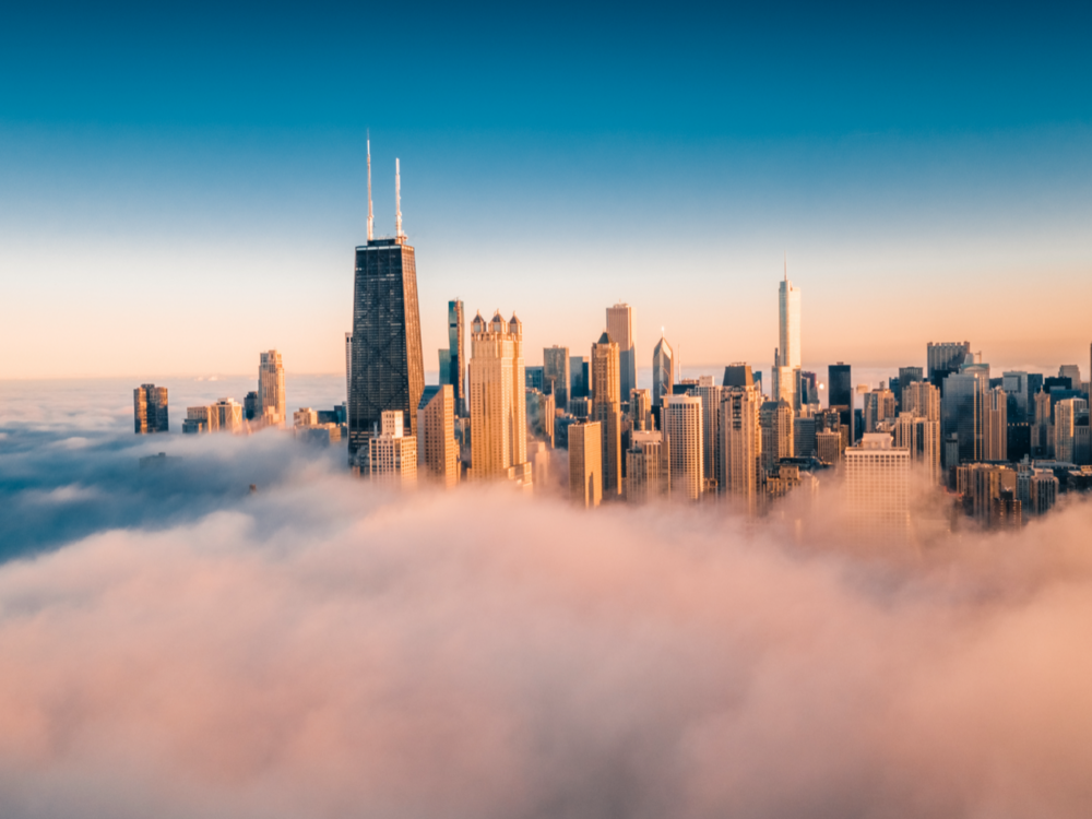 Aerial view of a city skyline with tall skyscrapers rising above a layer of thick clouds during sunrise, under a clear blue sky.