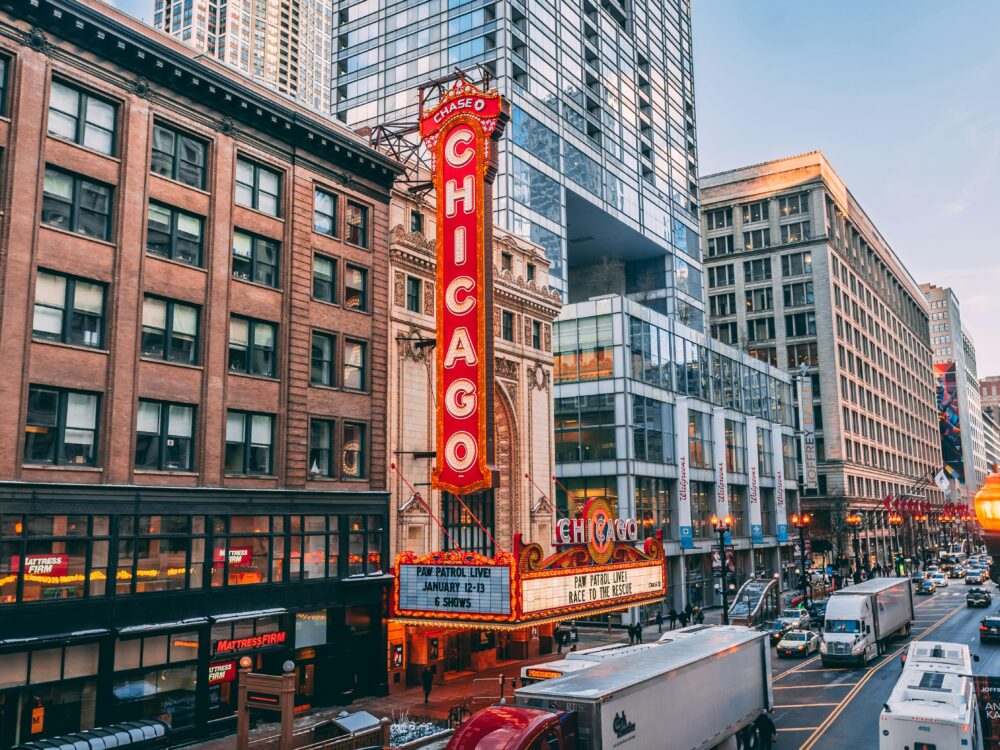 Downtown Chicago street scene with the iconic red Chicago Theatre sign, surrounded by historic and modern buildings, cars, and pedestrians under a clear sky.