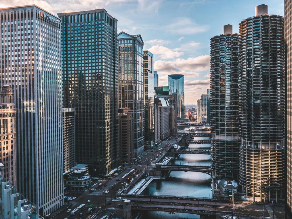 A cityscape view of downtown Chicago featuring tall skyscrapers, the Chicago River with bridges crossing it, and reflections of buildings in the water under a partly cloudy sky.
