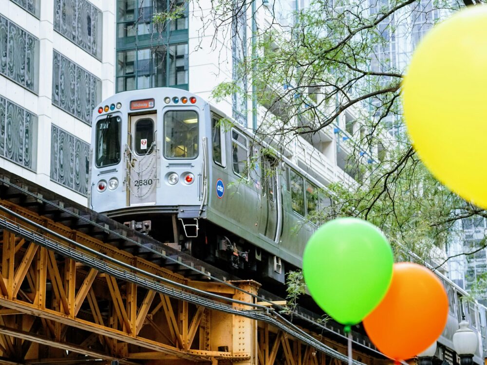 A silver elevated train passes through a city with modern buildings, while yellow, green, and orange balloons are in the foreground. Tree branches are visible near the tracks.