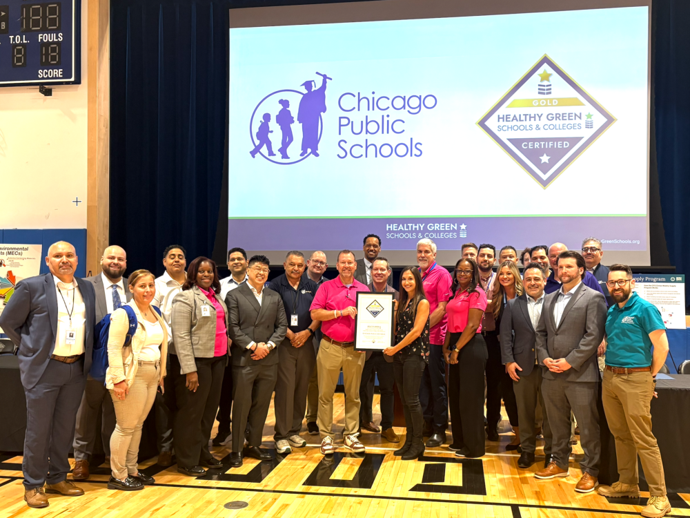 A group of people poses in a gym, holding a framed certificate, in front of a screen displaying the Chicago Public Schools logo and a Healthy Green Schools & Colleges Gold Certification badge.