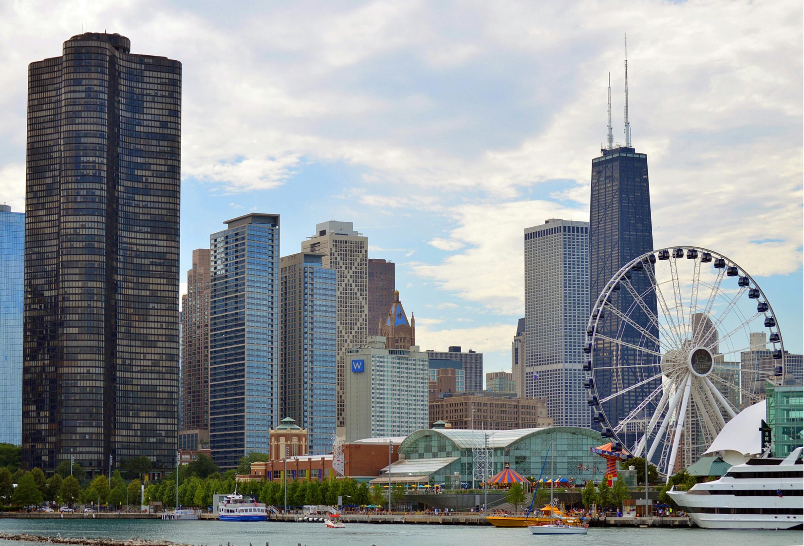 Chicago skyline with tall modern buildings, a large Ferris wheel, and boats along the waterfront at Navy Pier on a partly cloudy day.