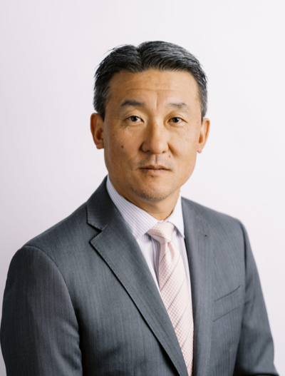 A man in a gray suit, light pink tie, and striped shirt poses against a plain light background. He has short, neatly styled dark hair and a neutral expression.