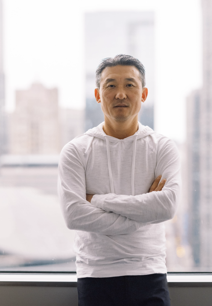 A man in a white hoodie stands indoors with his arms crossed, in front of a window overlooking a cityscape with tall, blurry buildings in the background.