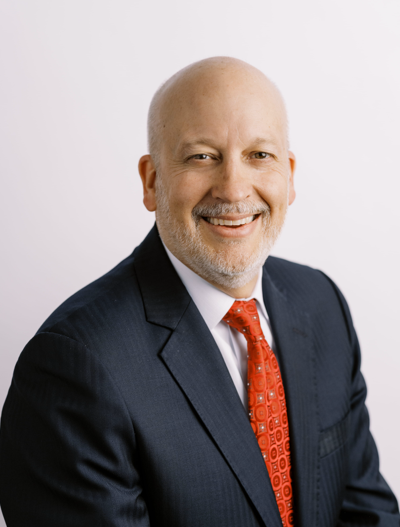 Smiling middle-aged man with a bald head and gray beard, wearing a dark suit, white shirt, and a red patterned tie, posing in front of a plain light background.