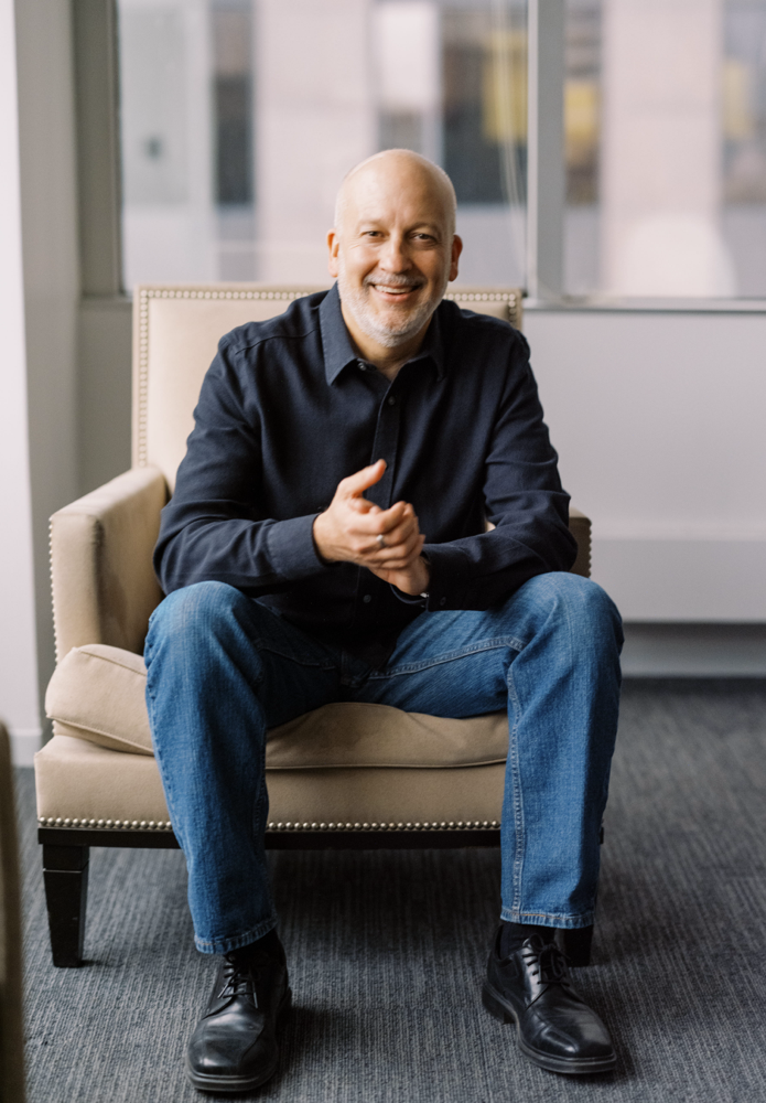 A bald man with a beard, wearing a dark shirt and blue jeans, sits and smiles in a beige armchair in an office with large windows.