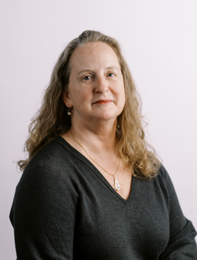 A woman with long wavy hair, wearing a dark V-neck sweater and a necklace, sits against a light pastel background, looking calmly at the camera.