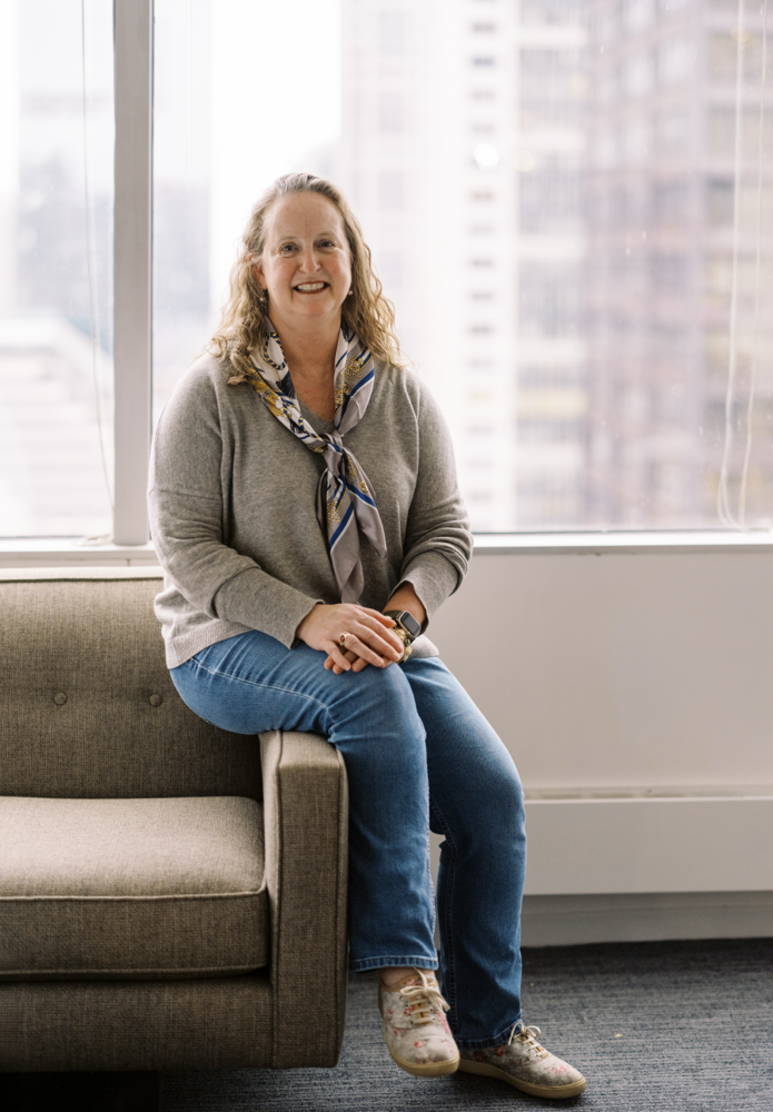 A woman with curly hair, glasses, and a scarf sits on the arm of a beige sofa in front of large windows overlooking tall city buildings. She is smiling and wearing a gray sweater, jeans, and floral sneakers.