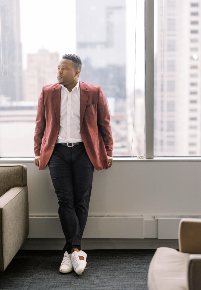A man in a red blazer, white shirt, and dark pants leans against a windowsill in a modern office with large city skyscrapers visible outside. He looks out the window with one foot crossed over the other.