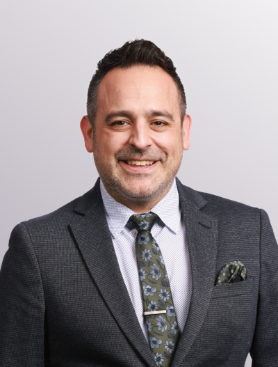 A man with short dark hair and a trimmed beard smiles at the camera. He is wearing a dark textured suit, a light collared shirt, and a floral patterned tie with a matching pocket square. The background is light gray.