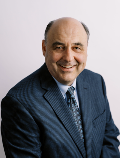 A middle-aged man with a receding hairline, wearing a blue patterned suit jacket, white shirt, and patterned tie, smiles while sitting against a plain light background.