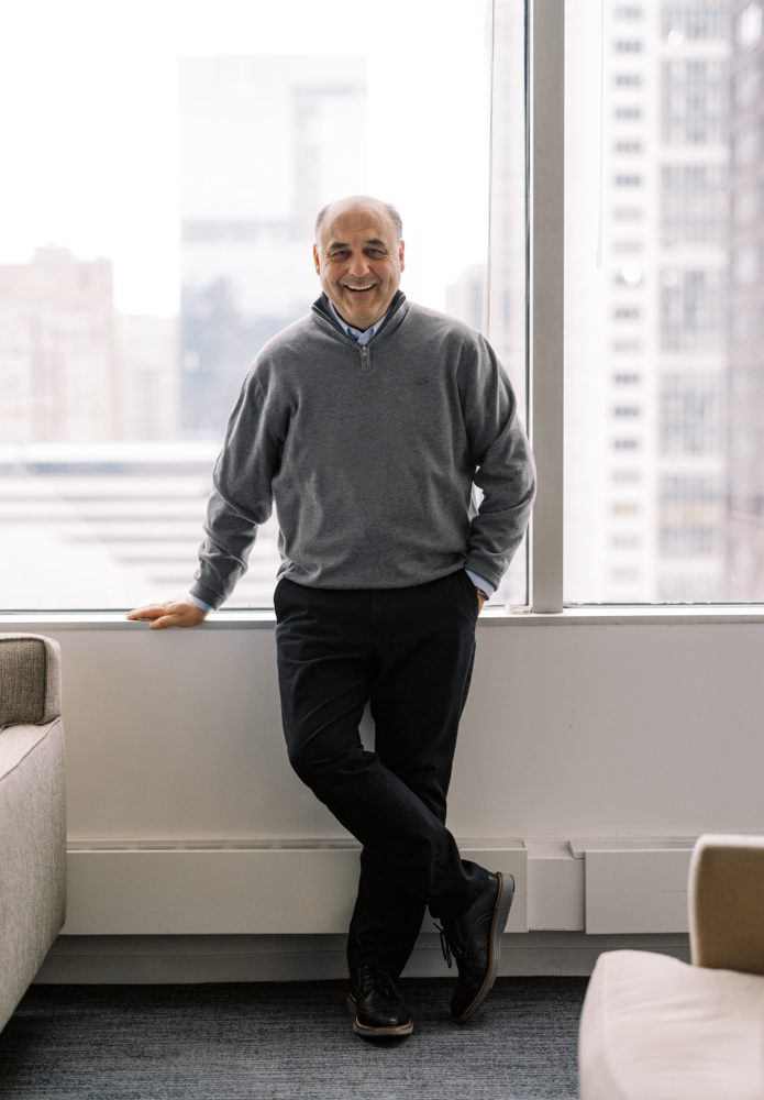 A man stands in a modern office by a large window with city buildings in the background, smiling and leaning slightly against the windowsill with his arms relaxed. He is dressed in a grey sweater and dark pants.