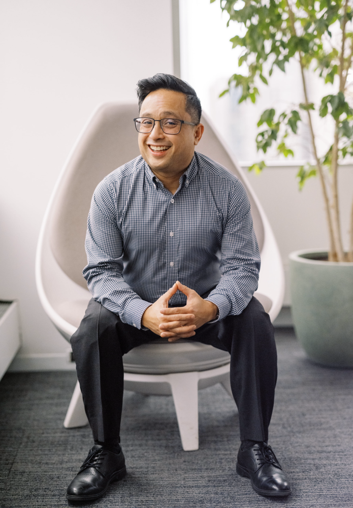 A person wearing glasses, a checkered shirt, and black pants sits smiling on a modern white chair in an office setting with a potted plant in the background.