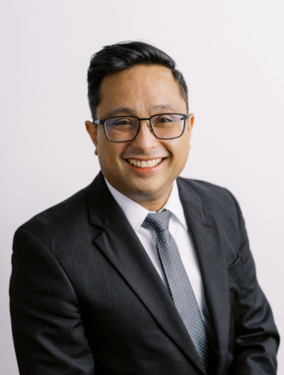 A man wearing glasses, a dark suit, a white shirt, and a dotted tie smiles at the camera against a plain light background.