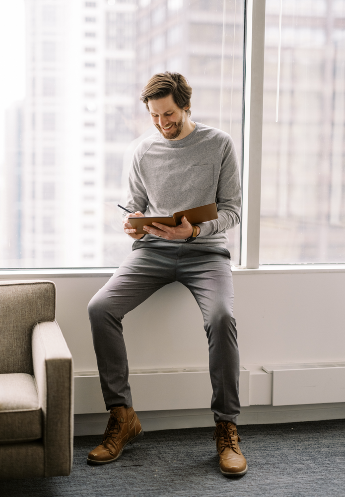 A man in a gray sweater and pants sits on a windowsill, writing in a notebook. He is smiling and looking down at the notebook. Tall buildings are visible through the large window behind him.