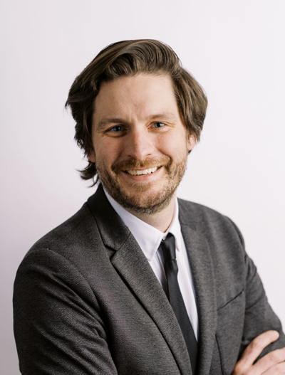 A man with medium-length brown hair and a beard, wearing a dark gray suit, white shirt, and black tie, smiles at the camera against a plain white background.
