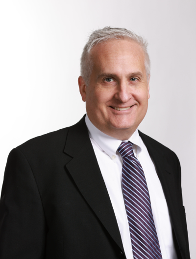 A middle-aged man with short gray hair, wearing a black suit jacket, white dress shirt, and striped tie, smiles at the camera against a plain white background.