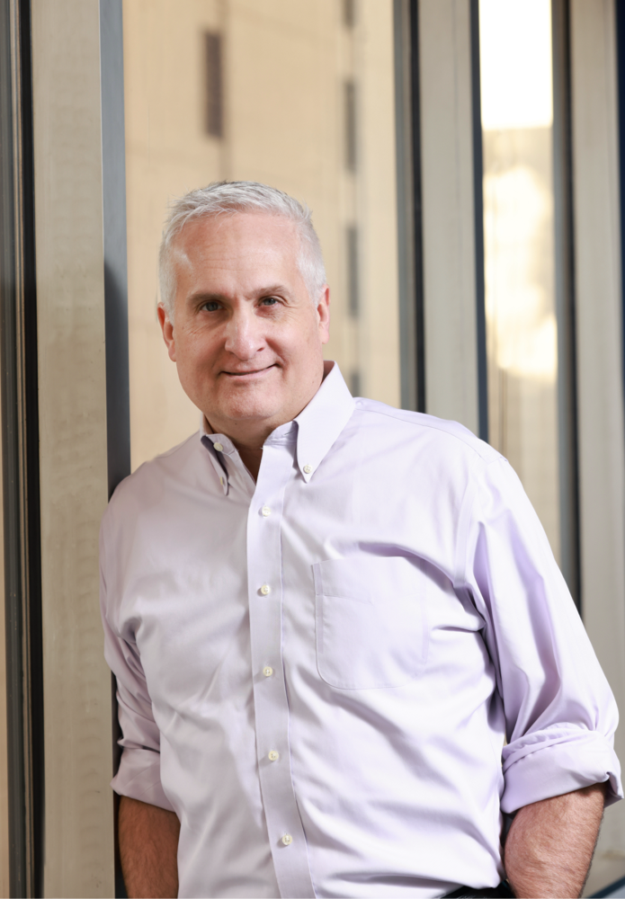 A middle-aged man with short gray hair, wearing a light purple button-down shirt with rolled-up sleeves, stands indoors near large windows, smiling slightly at the camera.