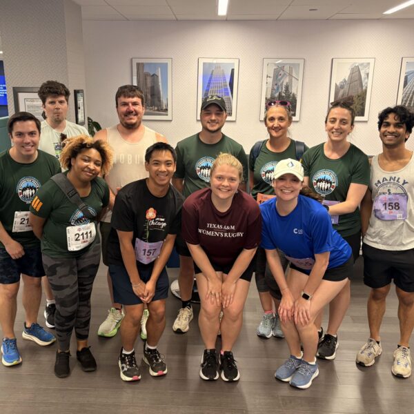 A group of twelve people in athletic wear pose and smile for a photo indoors, some with race bibs on their shirts. The setting appears to be a lounge area with couches, tables, and framed photos on the wall behind them.