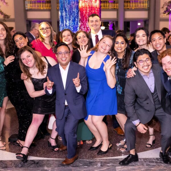 A group of people dressed in colorful formal attire pose cheerfully together at an indoor event with festive lights and decorations in the background. Most are smiling, some holding up peace signs or waving at the camera.