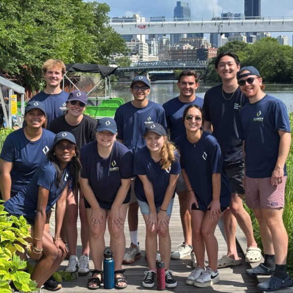 A group of 12 people in matching navy blue shirts and caps pose together on a wooden dock surrounded by lush greenery, with a city skyline and a bridge visible in the background on a sunny day.