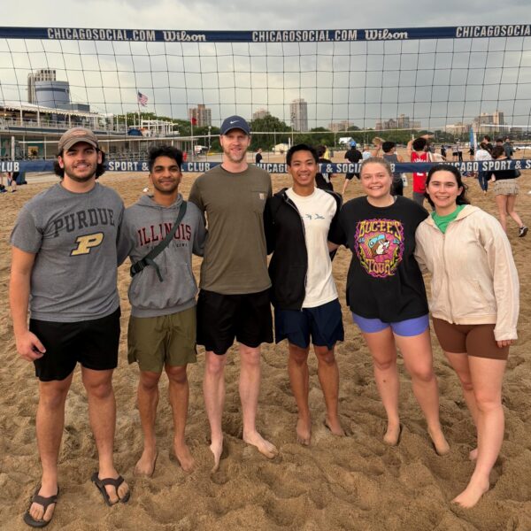 Six people stand on a sandy volleyball court, smiling at the camera. A volleyball net and cloudy sky are in the background. All are dressed casually, wearing shorts and T-shirts or sweatshirts.