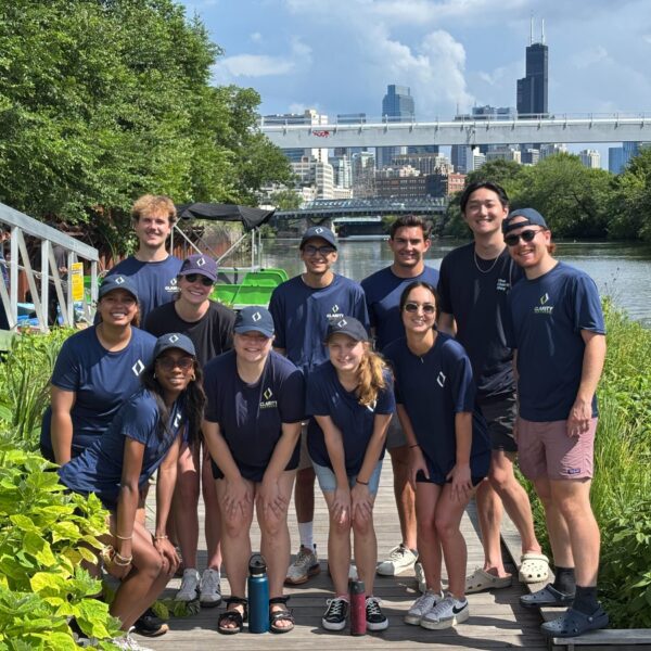 A group of 12 smiling young adults in matching navy shirts and shorts pose on a dock by a river, surrounded by greenery, with a city skyline and bridge in the background on a sunny day, celebrating the start of their internships together.