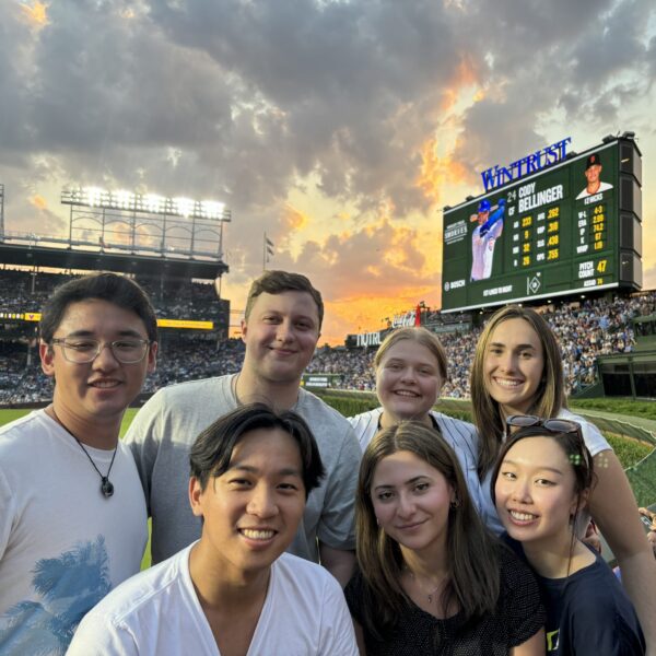 Seven young adults, gathered for internships, smile for a group photo at a baseball stadium during sunset. A large scoreboard and crowd are visible in the background under a colorful sky.