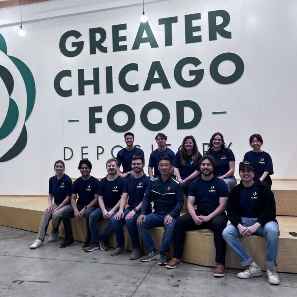 A group of people in matching shirts pose and smile in front of a large Greater Chicago Food Depository sign inside a spacious building with inspirational quotes on the wall above.